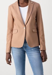 Blazer marron clair à texture lisse, fermeture à un bouton, deux poches avant et un emblème décoratif sur la poche gauche. Porté sur un haut blanc.