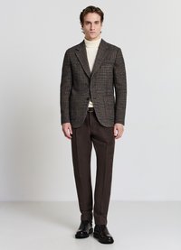 Blazer a quadri marroni sopra un dolcevita color crema, abbinato a pantaloni marroni scuri e stivaletti, su uno sfondo neutro.