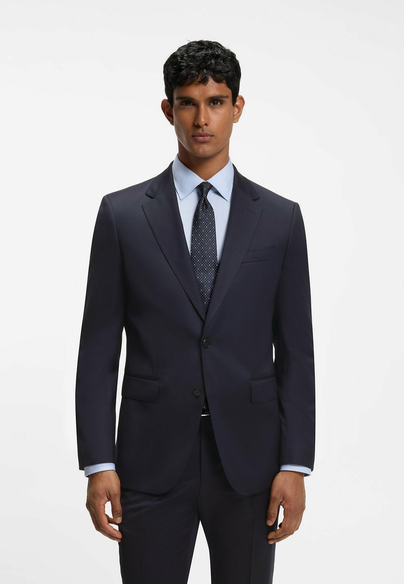 Homme aux cheveux courts et foncés portant un costume bleu marine foncé, une chemise bleu clair et une cravate foncée à motifs, debout devant un fond blanc uni.