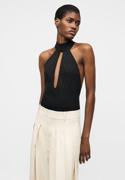 Femme aux cheveux courts portant un top noir sans manches à col bénitier avec une fente à l'avant et un pantalon blanc plissé taille haute à jambes larges.