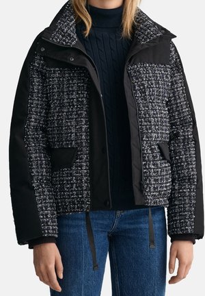 Vrouw draagt een zwart-wit geruit gewatteerd jack met effen zwarte panelen en blauwe jeans, staand met ontspannen armen langs het lichaam.