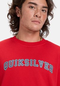 Červená mikina vyrobená z bavlny, na prednej strane má modrý nápis "QUIKSILVER" v oblúku. Textília má hladkú a rovnomernú textúru.