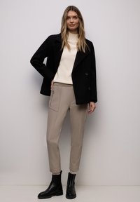 Frau steht vor weißer Wand, trägt eine schwarze Jacke, einen cremefarbenen Pullover, beige Hosen mit Reißverschlusstasche und schwarze Stiefeletten.