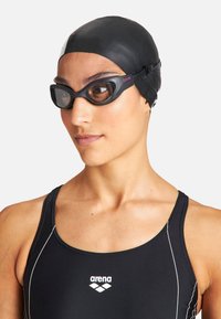 Gorro de natación negro, gafas de natación oscuras con lentes grises y una correa texturizada, traje de baño negro con detalles y logotipo blancos. Vista en primer plano.