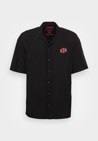 Maglietta nera a maniche corte con abbottonatura frontale, colletto classico e logo circolare rosso sul lato sinistro del petto. Tessuto con una texture liscia.