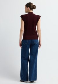 Top sin mangas burdeos con acentos de hombros con volantes, combinado con jeans de mezclilla azul de pierna ancha. La modelo está de espaldas a la cámara.
