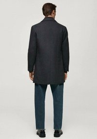 Dunkelgrauer Wollmischungs-Mantel mit taillierter Passform, mit Reverskragen und einer Schlitze, getragen über blauen Jeans und schwarzen Schuhen.