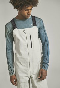 Weiße Latzhose mit verstellbaren schwarzen Trägern und Reißverschlusstaschen, kombiniert mit einem blauen Langarmshirt mit strukturiertem Muster und kontrastierenden Nähten.