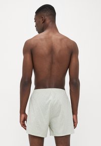 Mann med mørk hud og kort hår står med ryggen til, iført lysegrønne og hvite rutete shorts mot en enkel hvit bakgrunn.