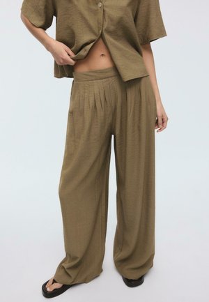 Person trägt locker sitzende, hochtaillierte olivgrüne plissierte Hose und dazu passendes, geknöpftes Hemd, steht vor einem einfarbigen hellen Hintergrund.