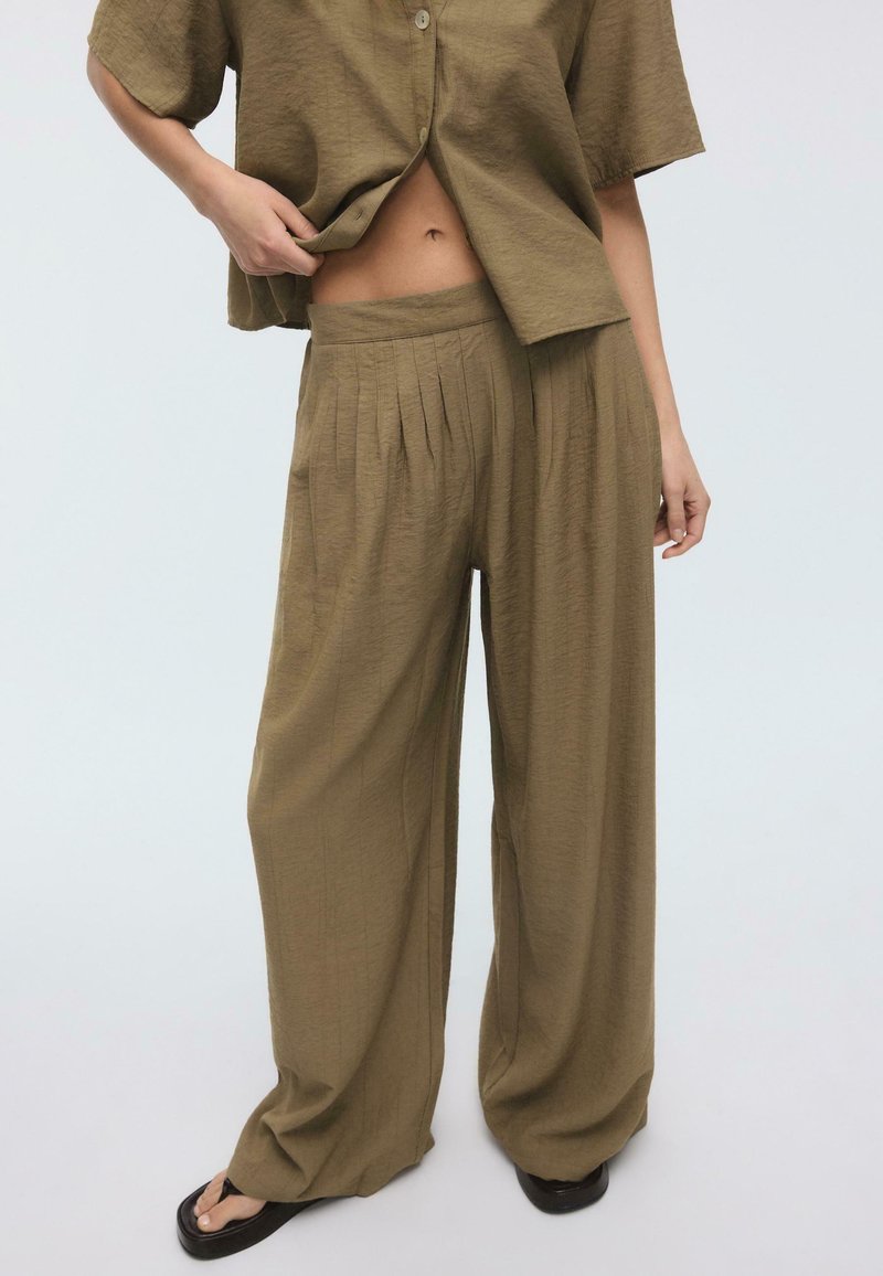 Personne portant un pantalon ample taille haute plissé vert olive et une chemise boutonnée assortie, debout devant un fond clair uni.
