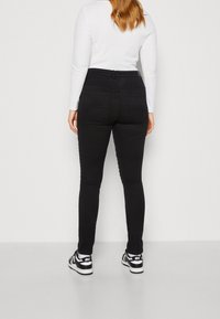 Vaqueros pitillo negros de cintura alta, con bolsillos traseros y textura suave, combinados con una camiseta blanca de manga larga y zapatillas negras.