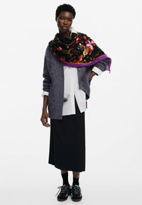 Cárdigan gris de punto trenzado sobre una camisa blanca, combinado con una falda negra. Una bufanda floral vibrante con ribete púrpura está drapeada alrededor del cuello. Zapatillas negras.