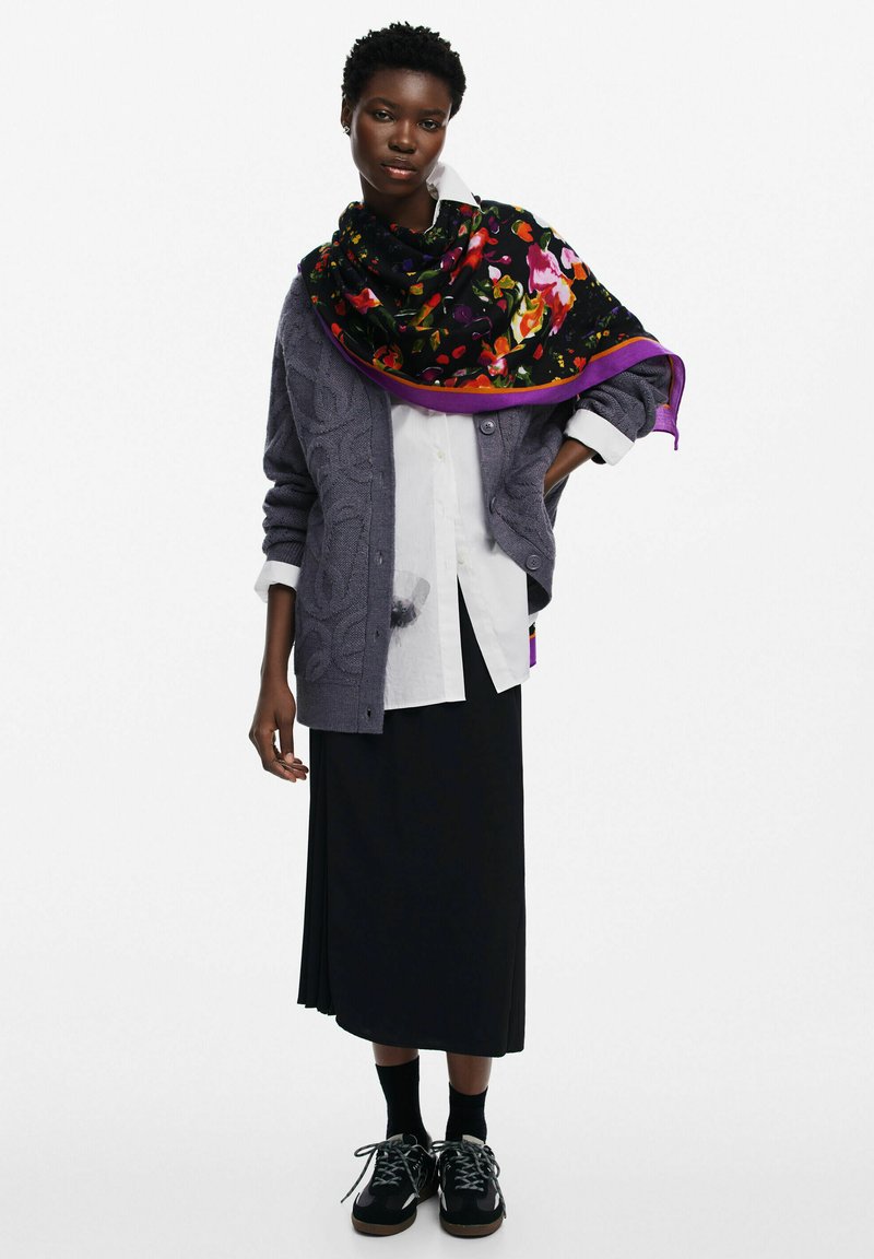 Cárdigan gris de punto trenzado sobre una camisa blanca, combinado con una falda negra. Una bufanda floral vibrante con ribete púrpura está drapeada alrededor del cuello. Zapatillas negras.