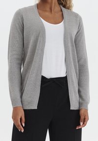 Šedý cardigan vyrobený z měkkého pleteného materiálu, s otevřeným předním dílem, dlouhými rukávy a žebrovanými manžetami, nošený přes bílý top a černé kalhoty.