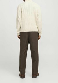 Personne debout, vue de dos, portant un pull en maille torsadée crème, un pantalon marron foncé et des chaussures marron, sur un fond uni.