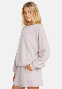 Light pink oversized sweatshirt with long sleeves, crew neck, and embroidered logo on the chest. Soft texture, relaxed fit, paired with matching shorts.