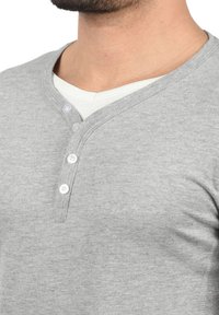 Šedé tričko typu henley s výstřihem do V, třemi bílými knoflíky a jemnou texturou. Pod ním je bílé tričko s kulatým výstřihem.