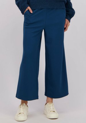 Brede broekspijpen in een rijke blauwe stof met een gladde textuur, voorzien van zijzakken en een ontspannen pasvorm, gecombineerd met witte sneakers.