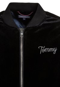 Czarna welurowa kurtka bomberka z srebrnym zamkiem, prążkowanym kołnierzem oraz wyszywanym białym logo "Tommy". Gładka tekstura, elegancki design.