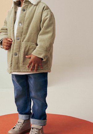 Kind draagt beige corduroy jas met sherpa kraag, opgerolde blauwe spijkerbroek en beige hoge sneakers, staand op een oranje ronde plaat.