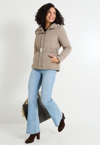 Chaqueta acolchada beige con cinturilla, gran cuello y botones a presión. La modelo lleva unos jeans acampanados de color azul claro y botas de tacón marrones.