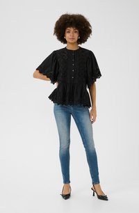 Schwarze Peplum-Bluse mit Spitzenverzierungen, kurzen Ärmeln und fünf Knöpfen. Kombiniert mit engen blauen Jeans und schwarzen High Heels.