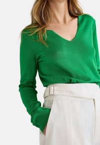 Pull en maille verte avec un col en V et des manches longues, associé à un pantalon blanc taille haute avec une boucle de ceinture argentée et des plis.