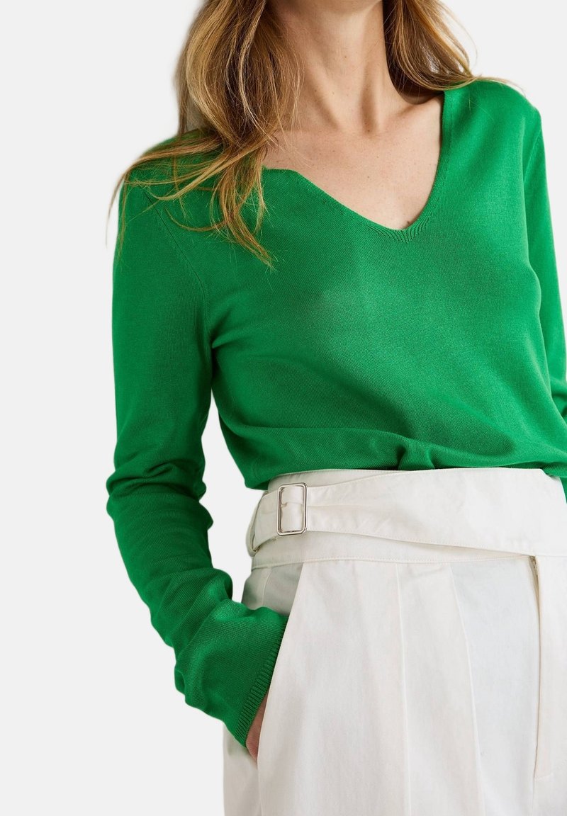 Pull en maille verte avec un col en V et des manches longues, associé à un pantalon blanc taille haute avec une boucle de ceinture argentée et des plis.