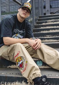 Svart grafisk t-shirt med logotyp, beige cargo-byxor med tigerbroderi, svarta sneakers och en färgglad hatt. Sitter på metalltrappor.