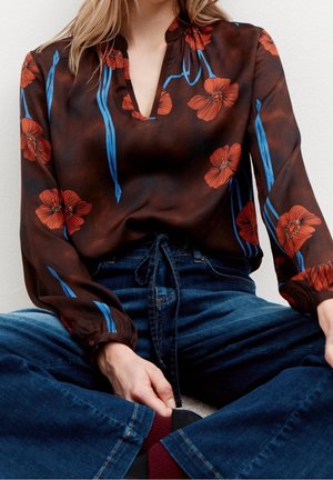 Femme portant un chemisier marron foncé avec des fleurs rouges et des tiges bleues, associée à un jean bleu taille haute, assise en tailleur.