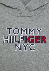 Szary materiał bluzy z kapturem z haftowanym tekstem "TOMMY HILFIGER NYC", podkreślającym "IGER" w czerwonym ściegu.