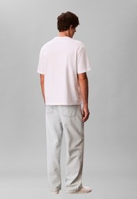 Άνδρας με την πλάτη γυρισμένη, φορώντας απλό λευκό μπλουζάκι, ανοιχτό γκρι τζιν και λευκά αθλητικά παπούτσια, με φόντο ουδέτερο γκρι.