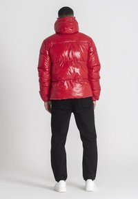 Rote glänzende Daunenjacke mit hohem Kragen, vertikalem Steppmuster, kombiniert mit schwarzen Hosen und weißen Sneakers, von hinten betrachtet.