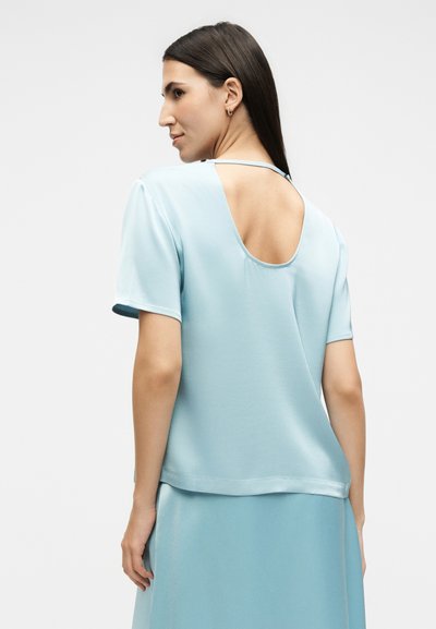 Γυναίκα με μακριά σκούρα μαλλιά που φοράει ένα ανοιχτό γαλάζιο τοπ με κοντά μανίκια και μεγάλη ανοιχτή πλάτη, καθώς και μια ταιριαστή φούστα, γυρισμένη προς τα πίσω σε απλό φόντο.