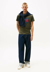 Camiseta verde oliva con logo rojo-azul, combinada con jeans de mezclilla oscuros de pierna ancha. Suéter azul marino colgado sobre los hombros, zapatillas blancas.