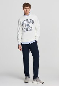 Felpa bianca con la scritta "NORTH DAKOTA COLLEGE" in stampa blu navale, abbinata a una camicia a polo azzurra e pantaloni scuri, completata da sneakers grigie.