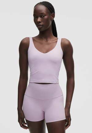 Kobieta z warkoczami, ubrana w liliowy, bezrękawnikowy crop top i pasujące szorty z wysokim stanem na jednolitym tle.