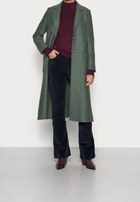 Zielony wełniany płaszcz o dopasowanym kroju, z dwoma przednimi kieszeniami i złotymi guzikami, noszony na bordowym golfie i ciemnoniebieskich sztruksowych spodniach.