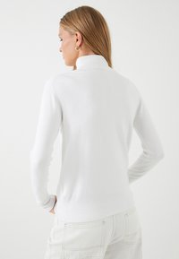 Vrouw met lang blond haar, gekleed in gouden creolen, een witte coltrui en witte broek, die met haar rug naar de kijker staat met haar handen op haar heupen.