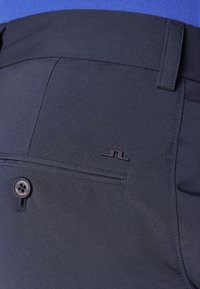 Marineblå bukser med glat stof, der har en baglomme med en knap og et lille sort logo på taljen.