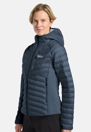 Vrouw in een marineblauwe gequiltte jas met capuchon, dichtgeritst, staand tegen een effen lichte achtergrond.