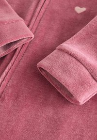 Suave tela de forro polar en rosa con puños doblados. Notable acento en forma de corazón en un tono más claro. Textura suave y detalle de cremallera visible.