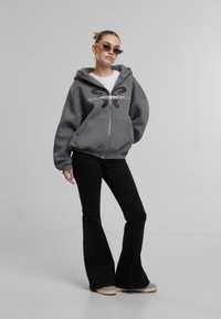 Grau zip-up Kapuzenpullover mit einem großen schwarzen grafischen Logo, kombiniert mit schwarzen Schlaghosen und grauen Schuhen, vor einem neutralen Hintergrund.