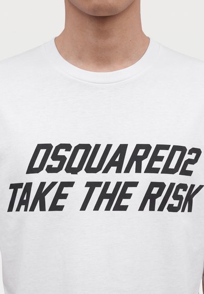 Homme portant un t-shirt blanc avec un texte noir "DSQUARED2 TAKE THE RISK" imprimé sur le devant.