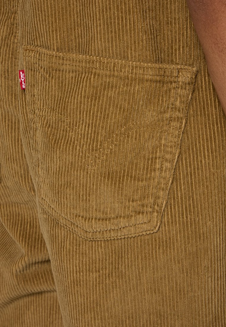 Tissu en velours côtelé marron avec des côtes verticales, comprenant une poche arrière et l'étiquette rouge de Levi's visible. La texture est douce avec un éclat subtil.