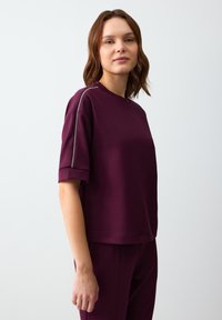 Bordeaux T-shirt met korte mouwen, gemaakt van zachte stof en met een ronde hals. Beschikt over zilveren accenten langs de schouders en heeft een relaxte pasvorm.