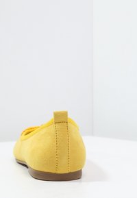 Žlutá z obecného zamaku plochá bota s poutkem na patě a hnědou podrážkou, umístěná na bílé ploše proti bílému pozadí.