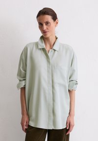 Light green oversized button-up shirt with a collar, single breast pocket, and rolled sleeves, made from a smooth fabric. Displayed on a model.