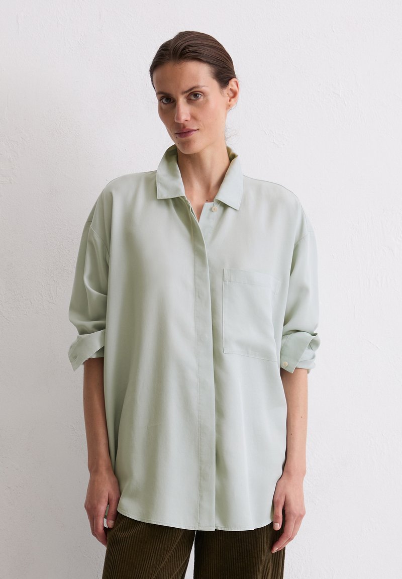 Light green oversized button-up shirt with a collar, single breast pocket, and rolled sleeves, made from a smooth fabric. Displayed on a model.